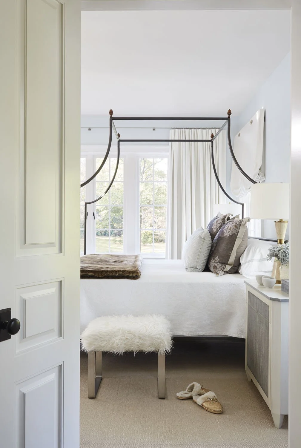 A bedroom with a four-poster bed, large window with white curtains, and a bedside table with a lamp. There is a furry stool and slippers on the beige carpet.