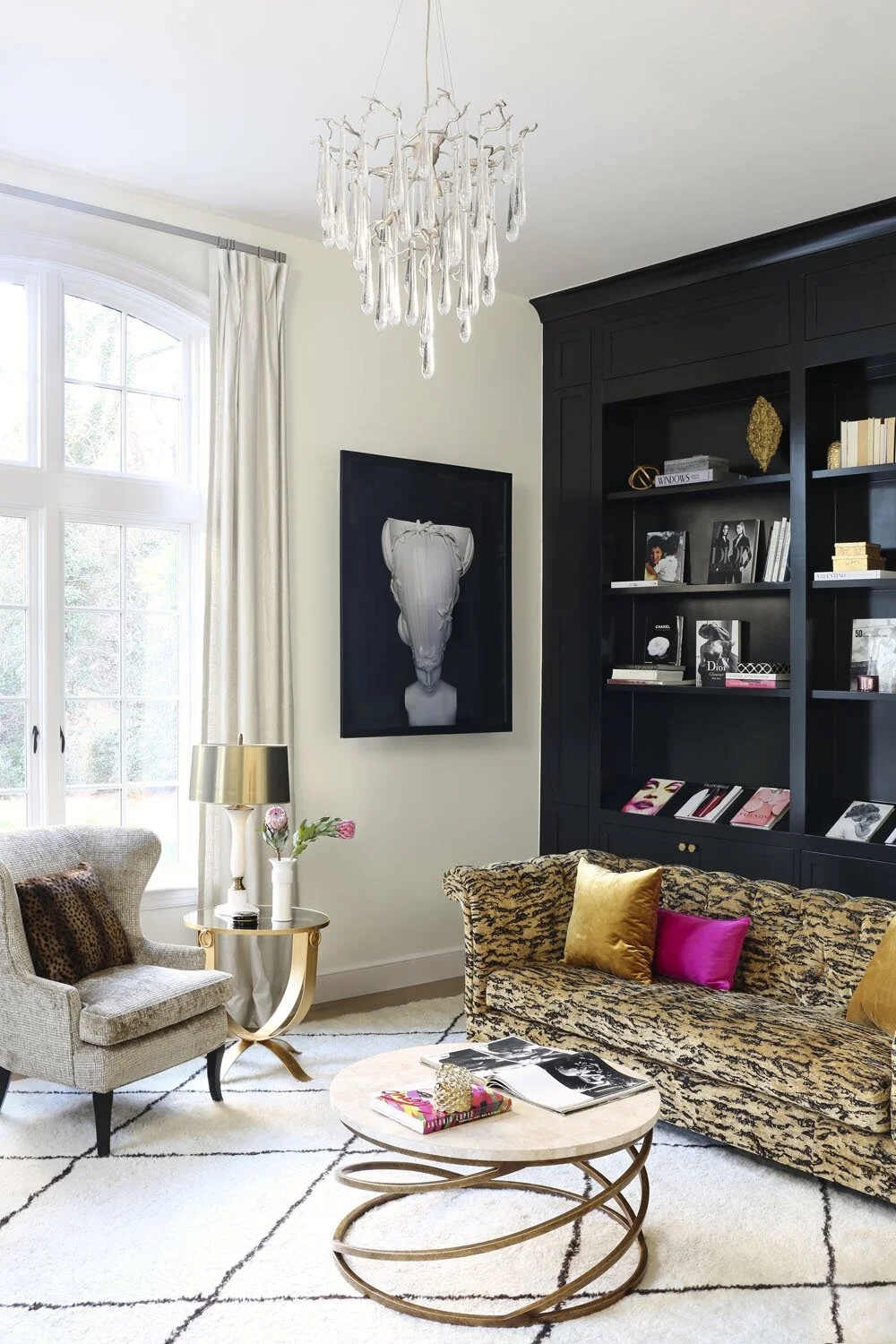 A living room with a large window, white curtains, a beige armchair, a black bookcase, a gold and black patterned sofa with colorful pillows, a small round coffee table with magazines, a tall table lamp, a chandelier, and decorative wall art.
