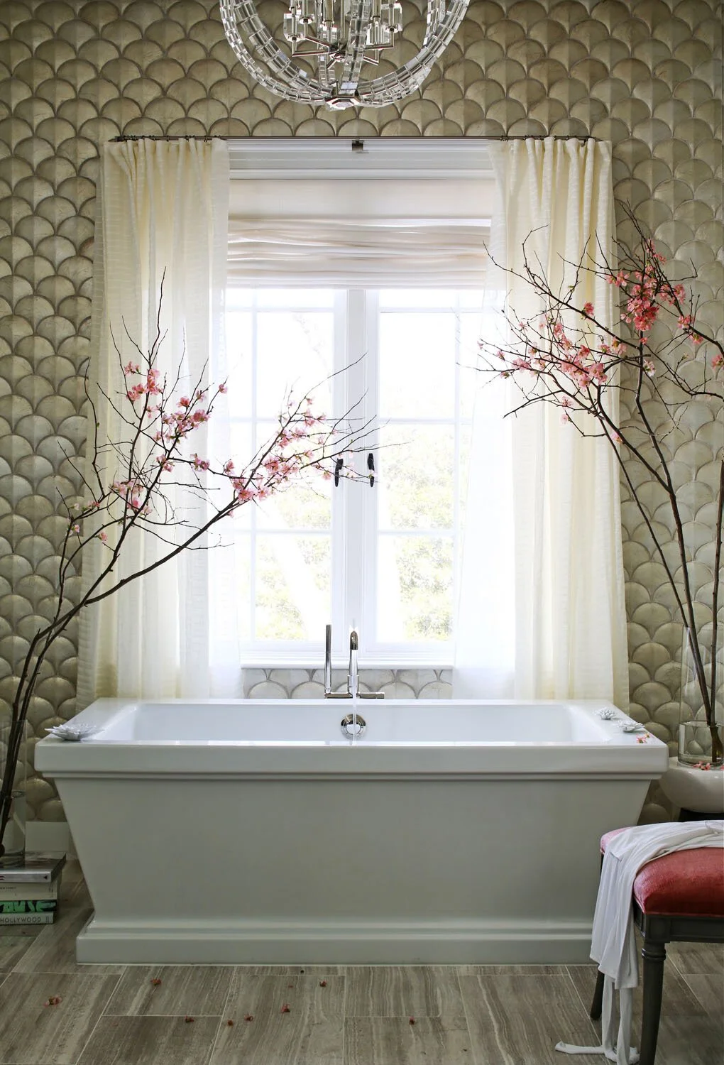 A bathroom with a white bathtub in front of a window, decorated with pink flowering branches on both sides and cream curtains. A chandelier hangs from the ceiling.
