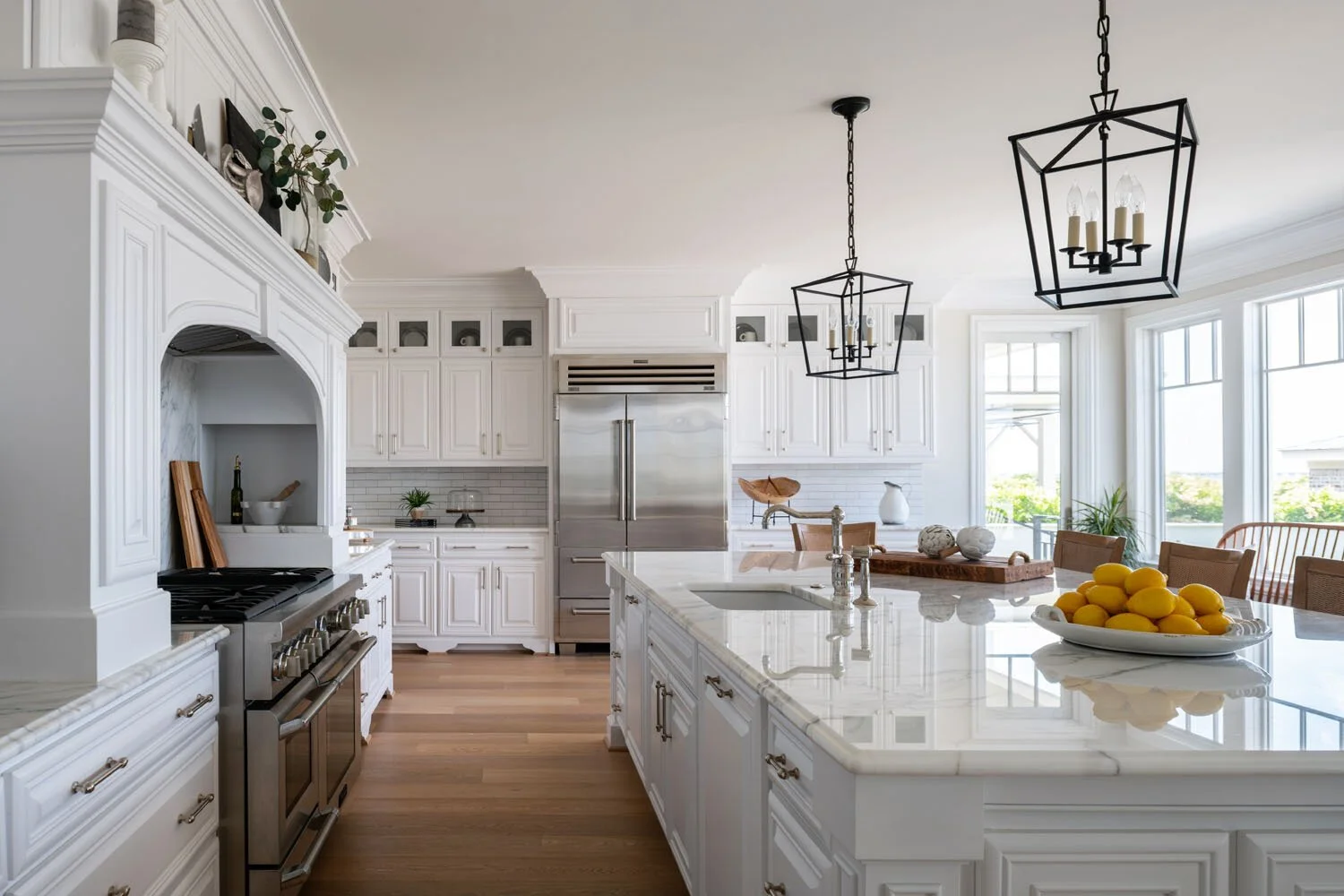 Bright kitchen with white cabinets, a large island with a marble countertop, stainless steel appliances, black geometric pendant lights, and large windows with a view of the outdoors.