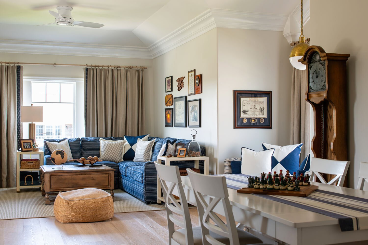 A living room with a large window, beige curtains, a blue striped sofa with navy and white pillows, a wooden coffee table, a white side table, wall art, and a dining table with white chairs and a chess set.