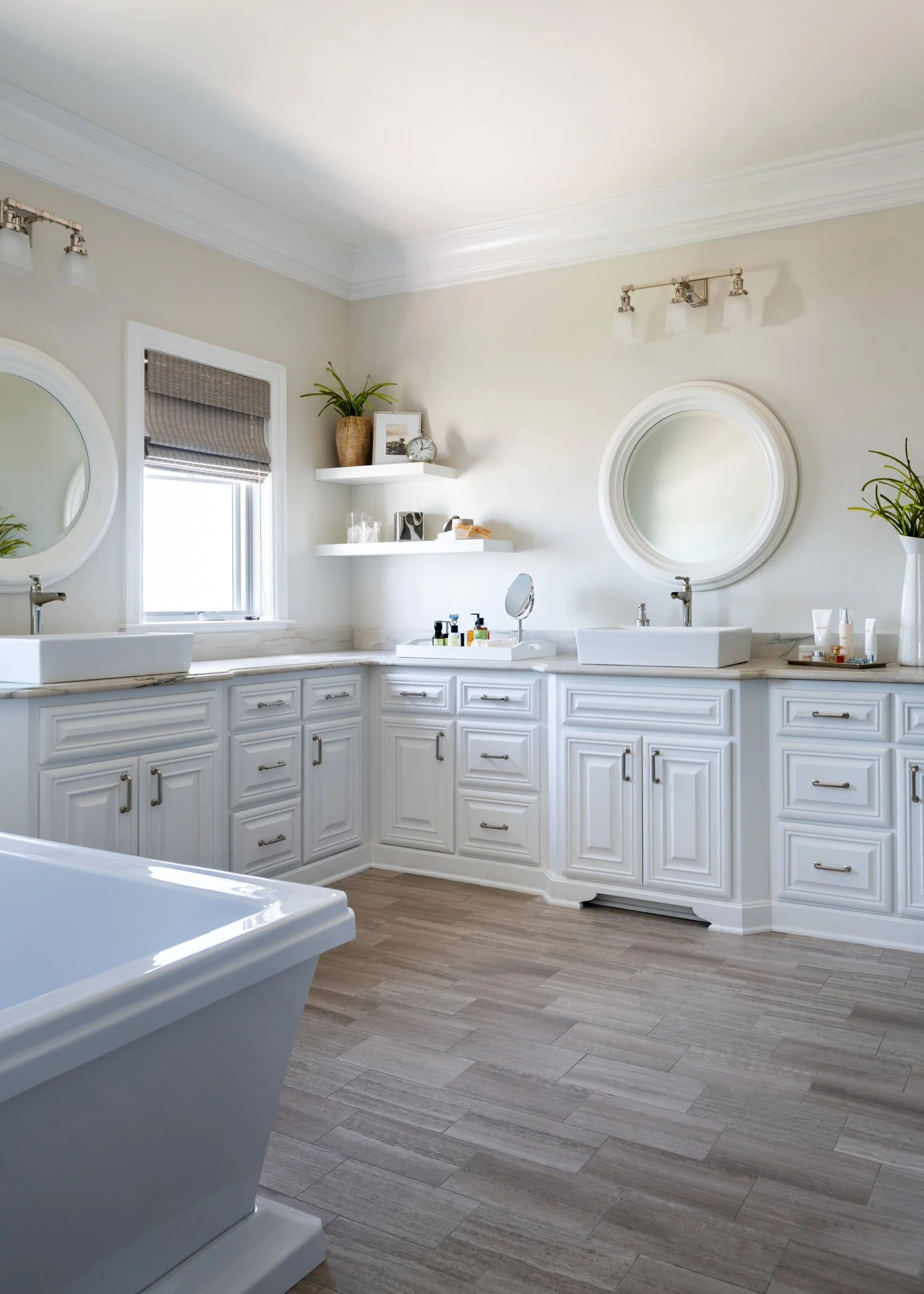 Bright bathroom with white cabinetry, oval mirrors above sinks, small window with a tissue box, potted plants, and shelves with decor items.