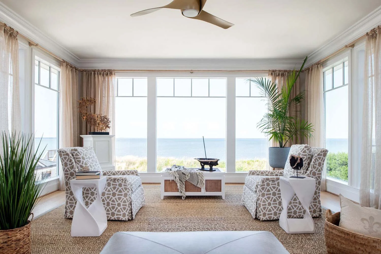 Living room with large windows overlooking the ocean, beige curtains, two patterned armchairs, green plants, a ceiling fan, and a woven rug.