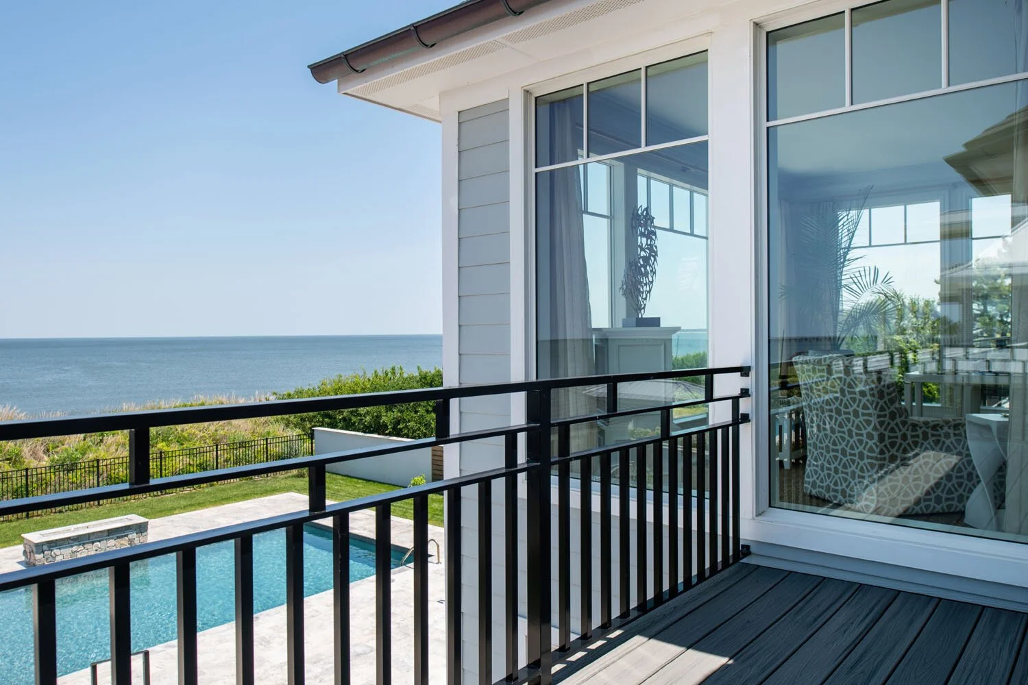 View from a balcony overlooking a swimming pool, grassy yard, and ocean in the distance, with a modern house interior visible through glass doors.