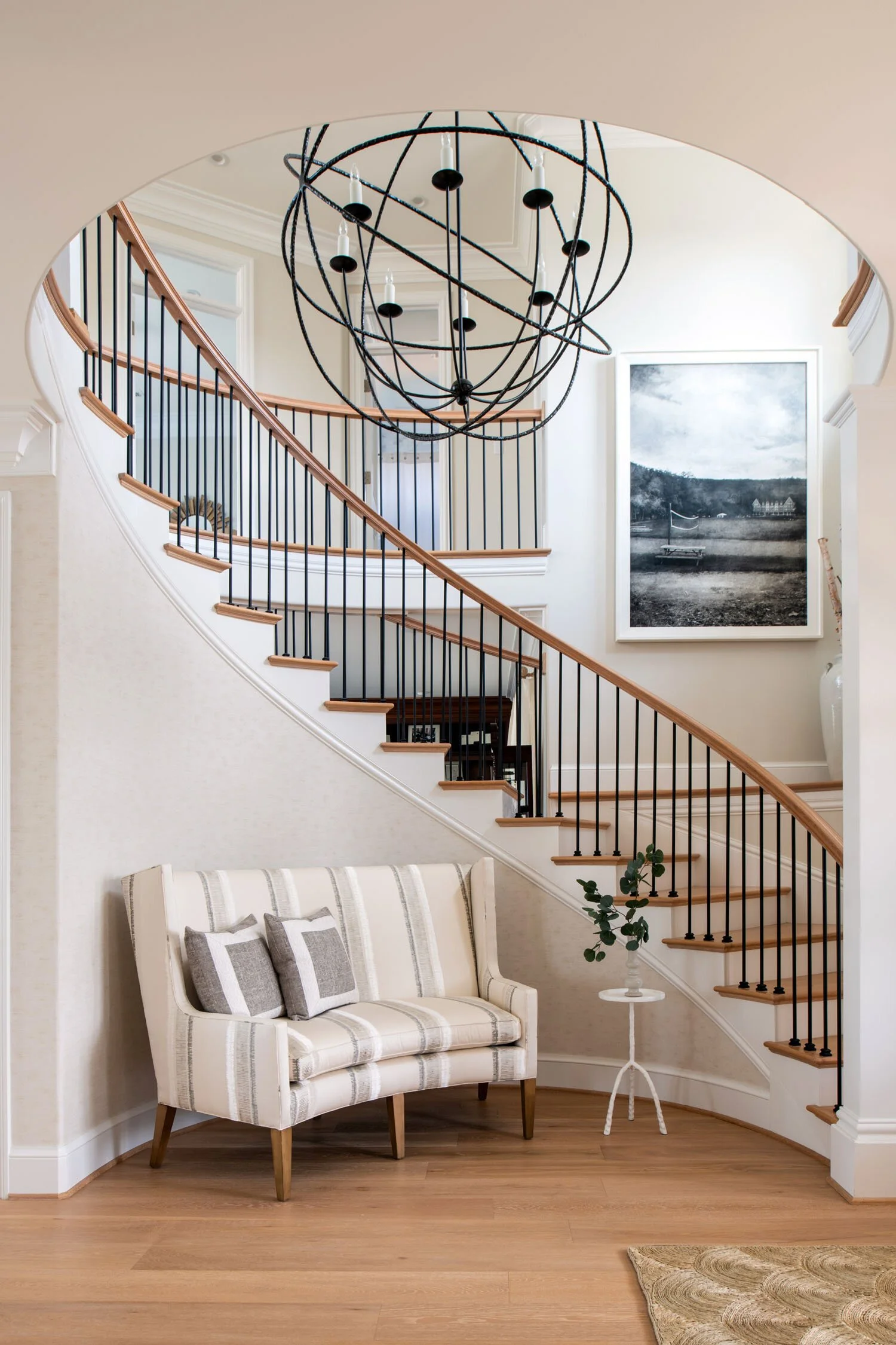 Interior view of a living space with a staircase featuring wooden treads and black metal balusters, a beige and white striped loveseat with two gray pillows, a small white side table with a green plant, a large black and white landscape photograph on