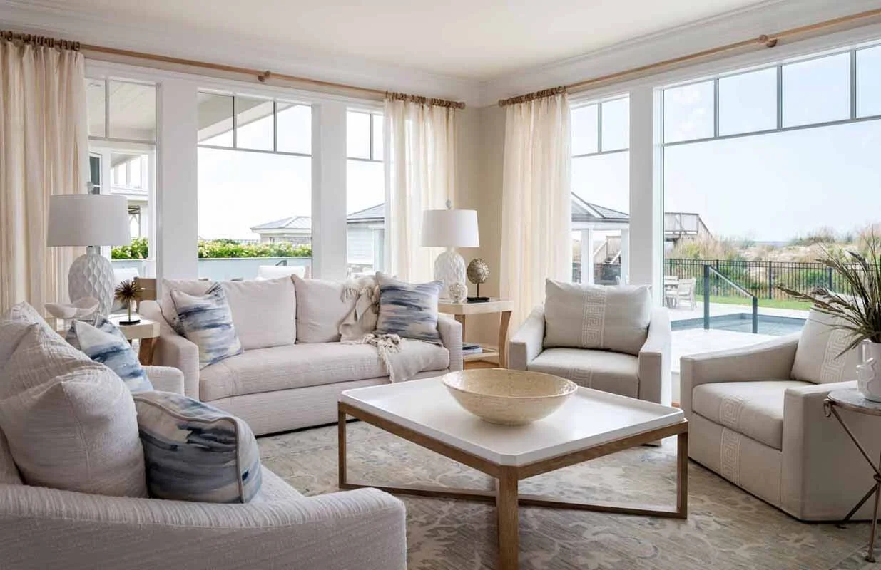Bright living room with white sofas, armchairs, and a coffee table. Large windows with cream curtains, outdoor view with a pool and patio.