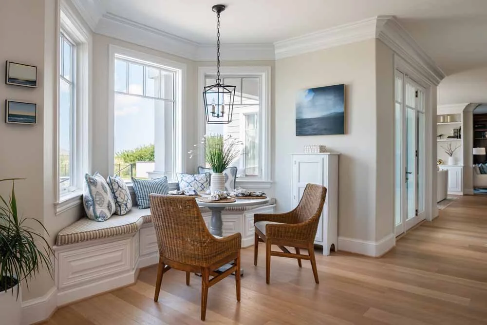 Bright dining nook with large windows, built-in bench with cushions, a small round table, two wicker chairs, and a hanging lantern light fixture.