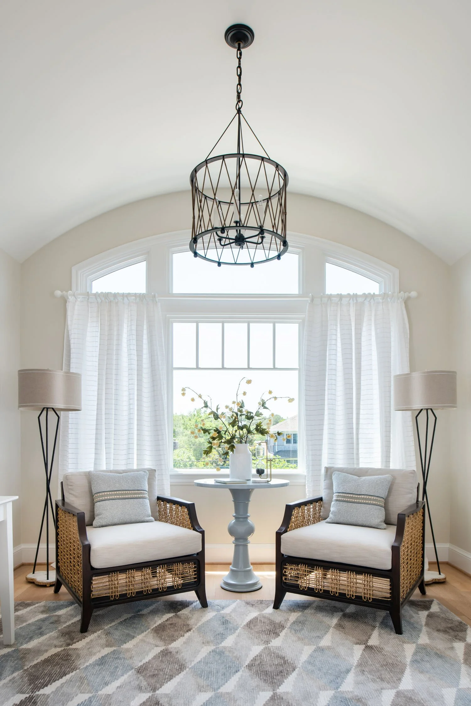 Living room with two armchairs, a round table in between, tall lamps, a window with white curtains, and a modern chandelier.