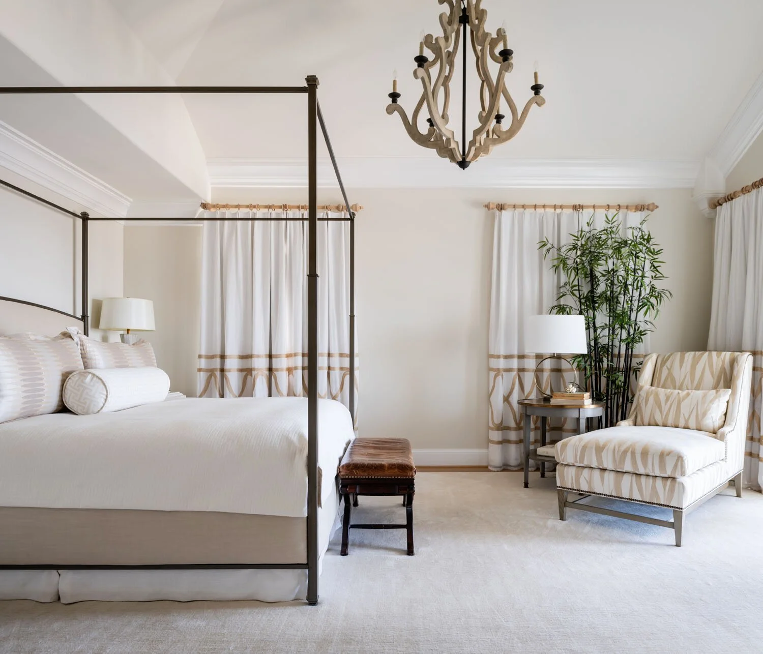 A well-decorated bedroom with a four-poster bed, a bench at the foot, a side table with a lamp and books, an armchair, a floor lamp, a large plant, and curtains, all in neutral tones.