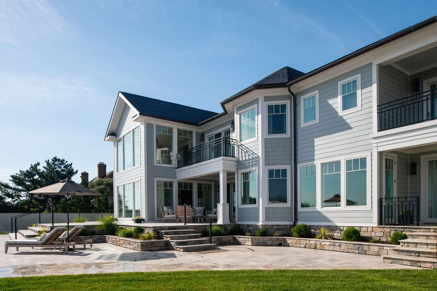 Front view of a modern, multi-story house with large windows, white siding, and a blue roof. There is a paved patio with lounge chairs, a small table with an umbrella, and landscaped plants and shrubs.