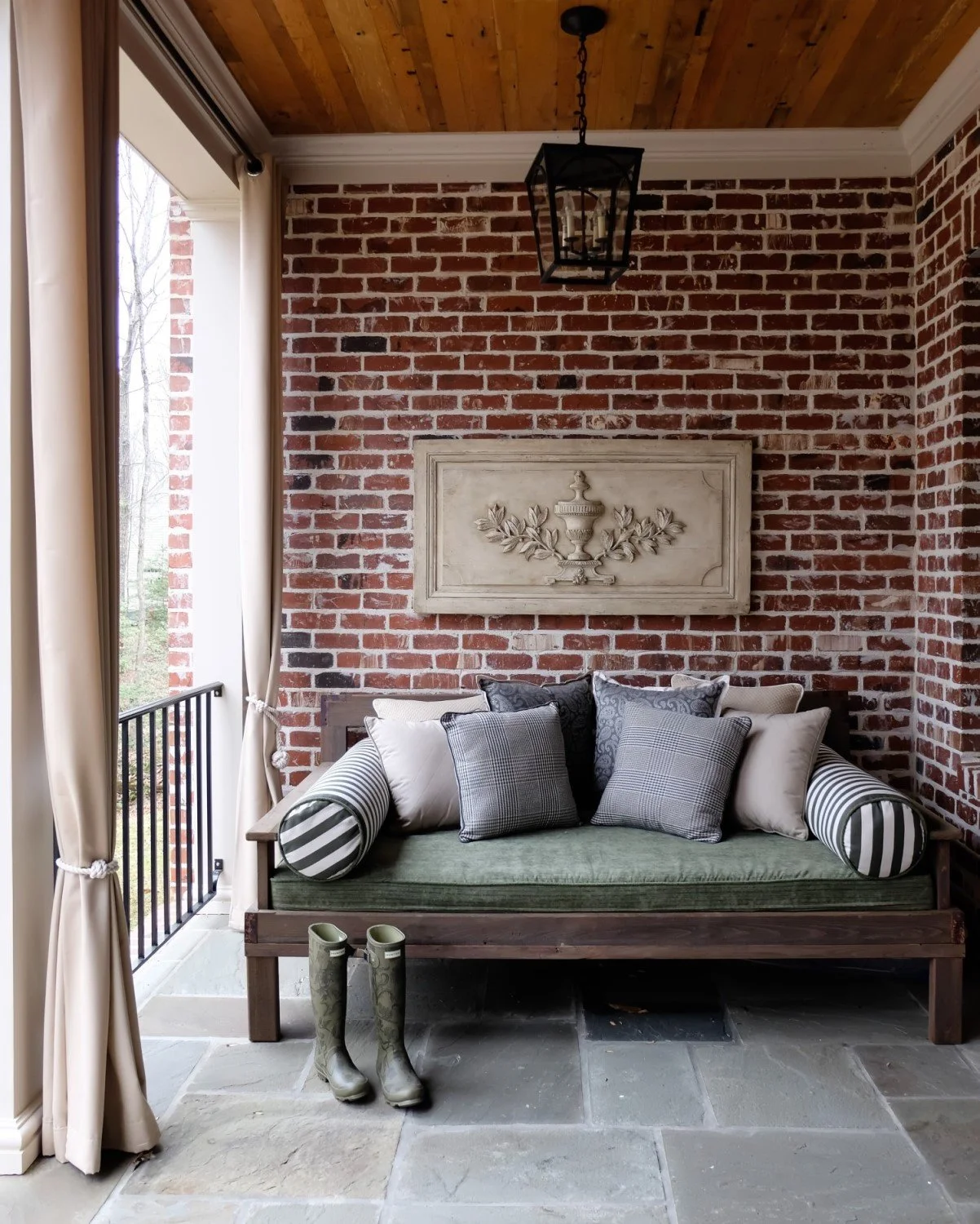 Living space with exposed brick wall, wooden ceiling, black lantern light fixture, decorative wall plaque, wooden bench with pillows, gray boots on stone floor near window with curtains.