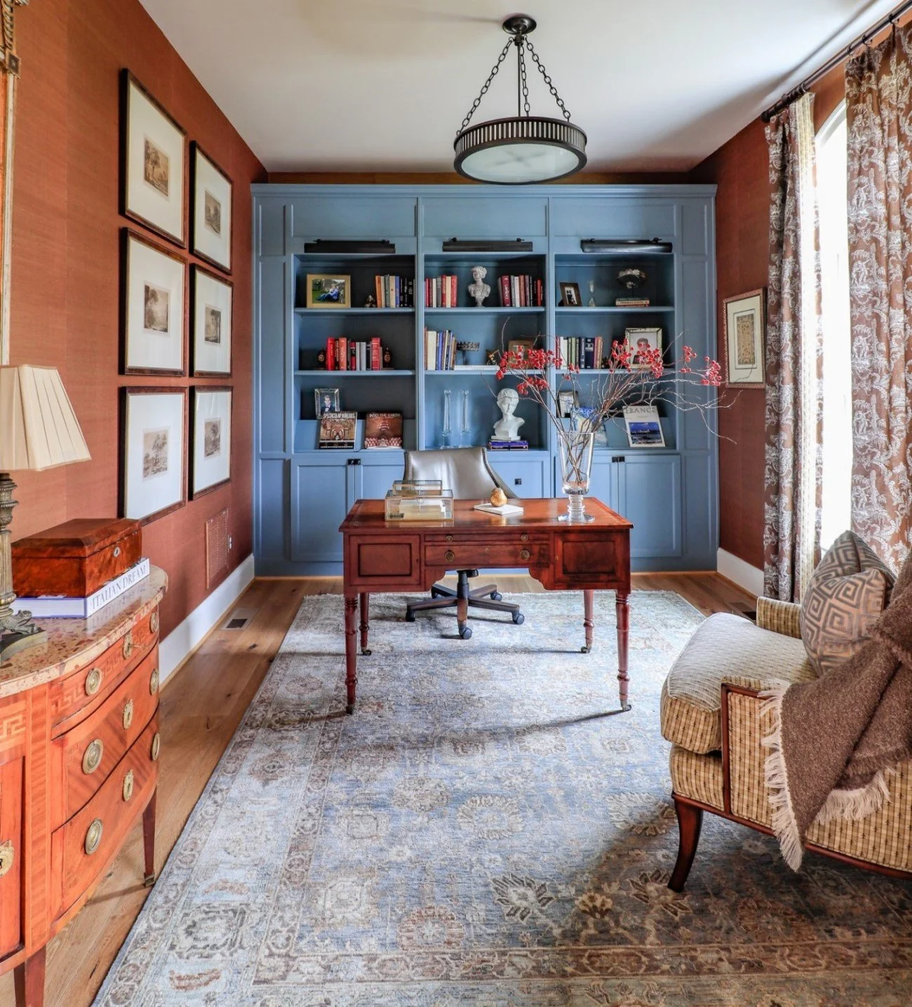 A cozy home office with a wooden desk, gray office chair, blue built-in bookshelves filled with books and decorative items, a patterned area rug, armchair with a blanket, large window with patterned curtains, framed artwork, and a ceiling light fixtu