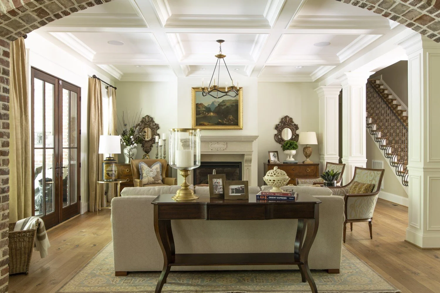 Elegant living room with beige sofa, armchairs, fireplace, framed landscape painting, chandelier, and large glass doors with gold curtains.