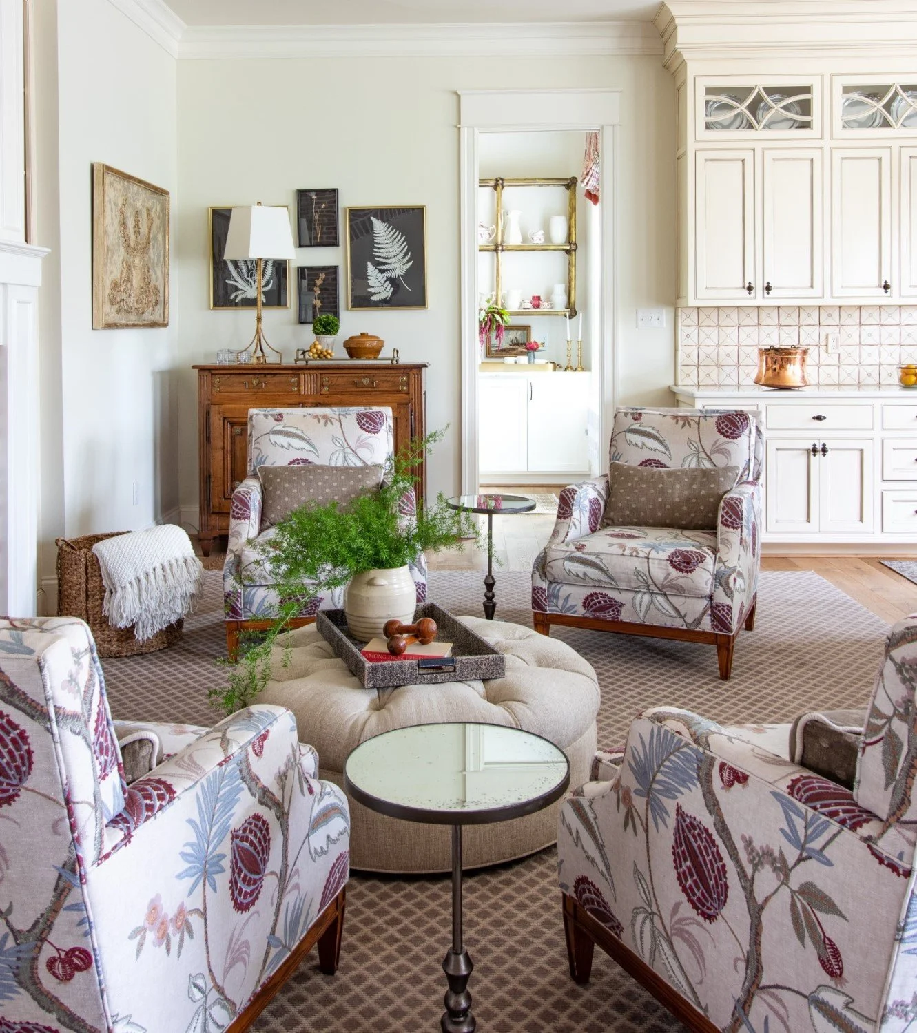 A cozy living room with patterned armchairs around a large tufted ottoman, a round side table, and a tray with decor. There's a wooden cabinet behind with framed artwork and a table lamp. An open doorway leads to a kitchen or pantry with shelves and 