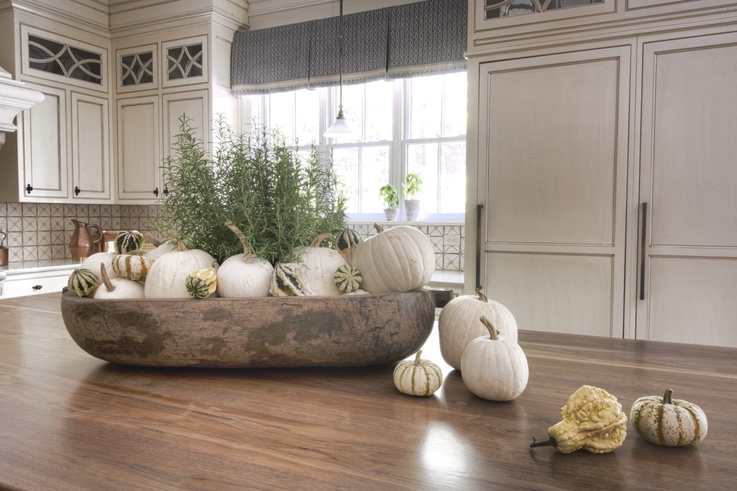 A large wooden bowl on a kitchen table filled with white gourds, decorative squash, and potted herbs, with a few gourds and squash outside the bowl on the table. The kitchen has white cabinetry, a tiled backsplash, and a window with blue patterned cu