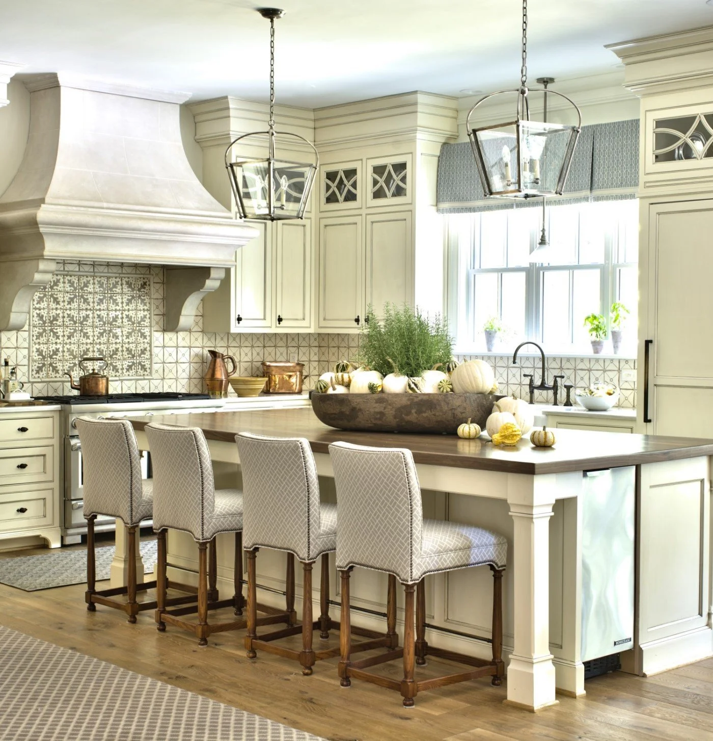 A bright, elegant kitchen with white cabinetry, a large center island with a wooden countertop, four upholstered bar stools, and decorative pumpkins and greenery on the island. Large windows with valances let in natural light.