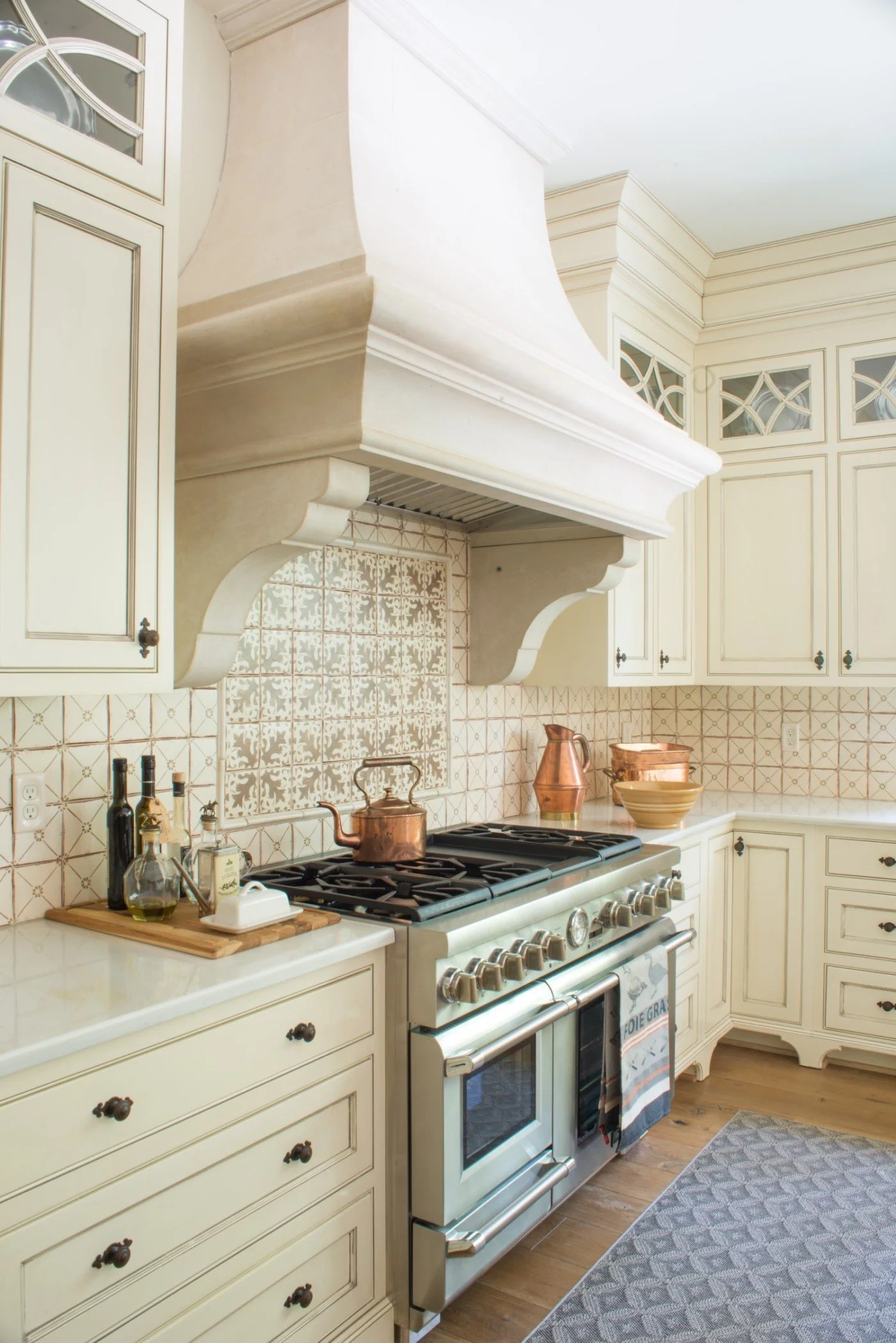 Cream-colored kitchen with a large stove, copper kettles, bowls, bottles, and a decorative tiled backsplash.