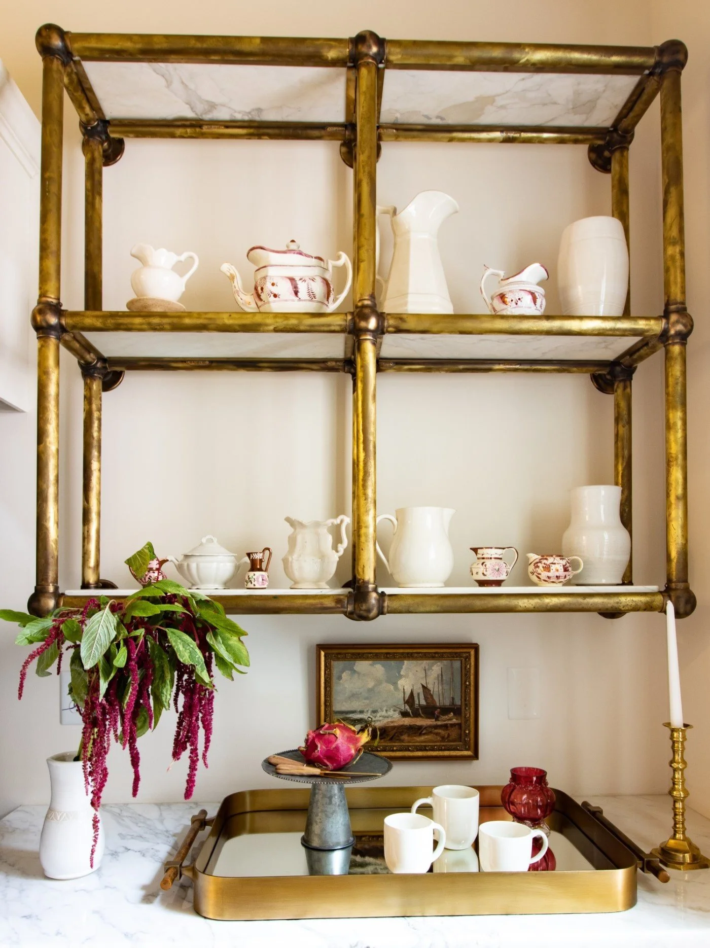 Decorative kitchen shelf with white ceramics, a pink flower, a framed painting, a white vase with pink flowers, and a gold candle holder with a white candle.