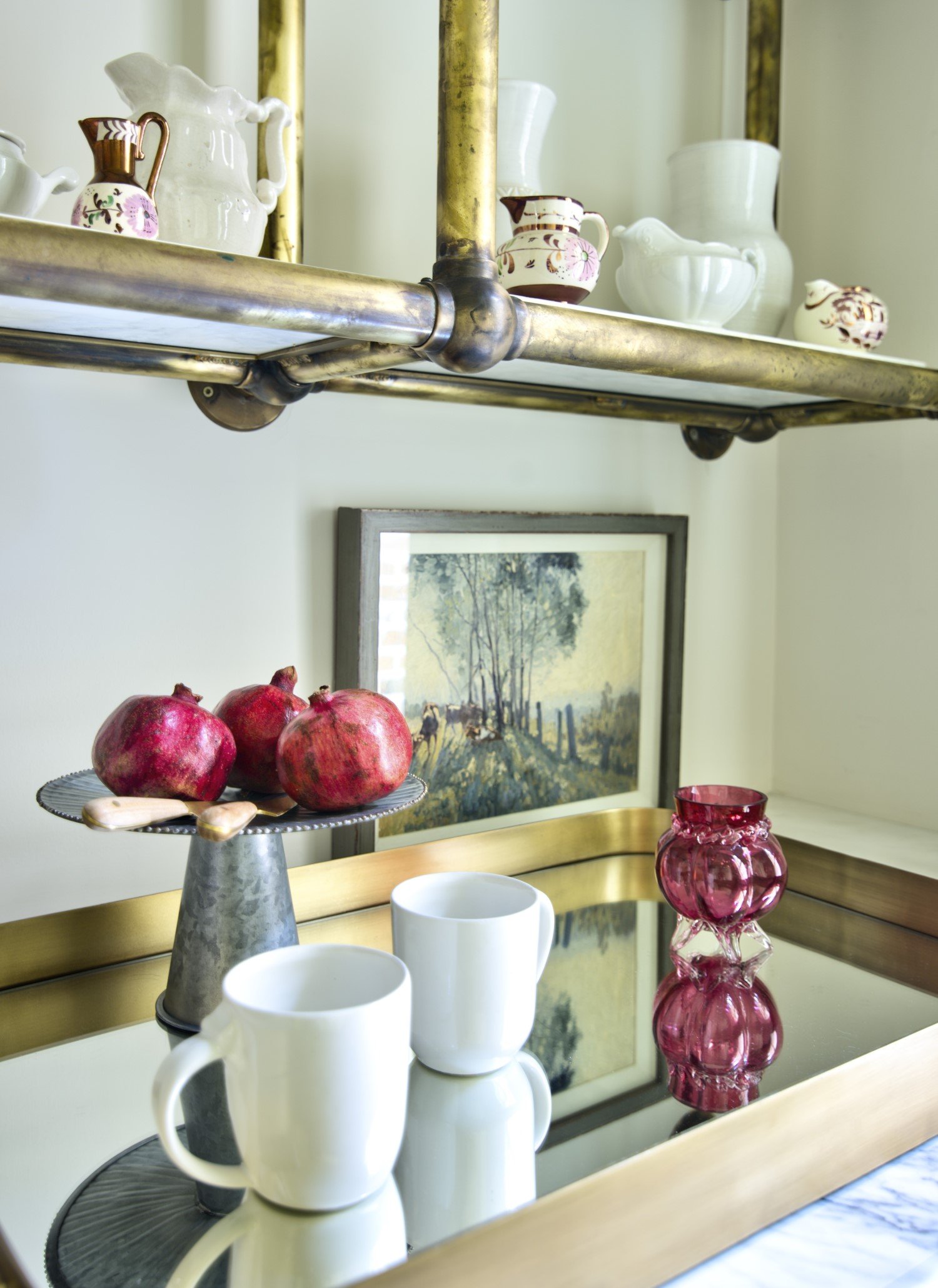 Close-up of a decorative table with two white coffee mugs, a pink glass vase, a pomegranate on a metal cake stand, and a framed landscape painting in the background. Stationery and a shelf with teapots and pitchers are visible above.