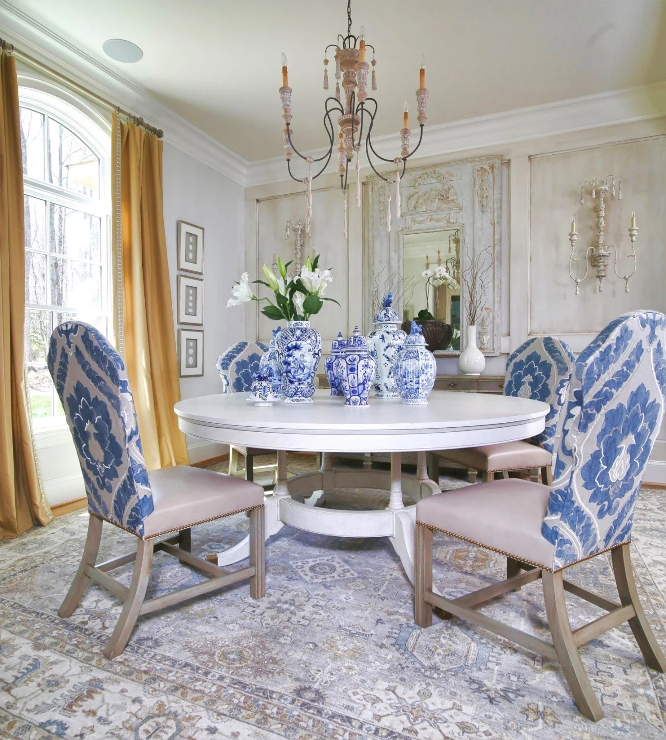 A dining room with an oval white table surrounded by six patterned blue and white chairs. The table has blue and white porcelain vases with flowers, a chandelier, and a mirror on the wall. Large windows with gold curtains and a patterned rug on the f