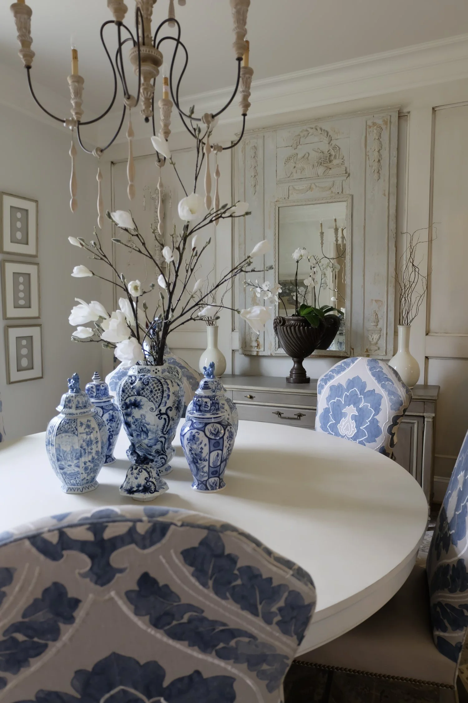 A dining room with a white oval table decorated with white and blue porcelain vases and a floral arrangement. The chairs have blue and white patterned upholstery. A chandelier hangs above, and there is a sideboard with large vases and a mirror on the