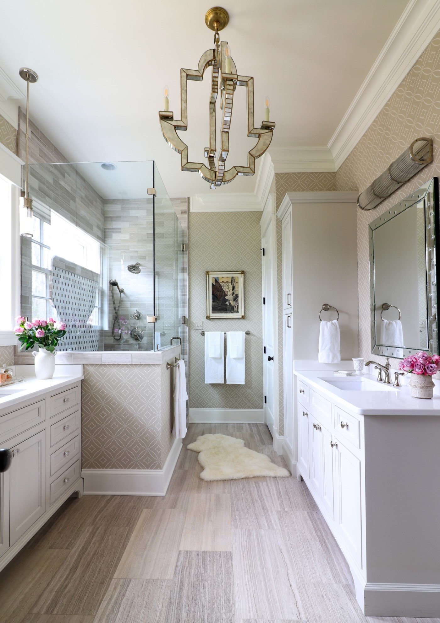 A bright, elegant bathroom with a chandelier, a glass-enclosed shower, and a double vanity with white cabinets and pink flowers.