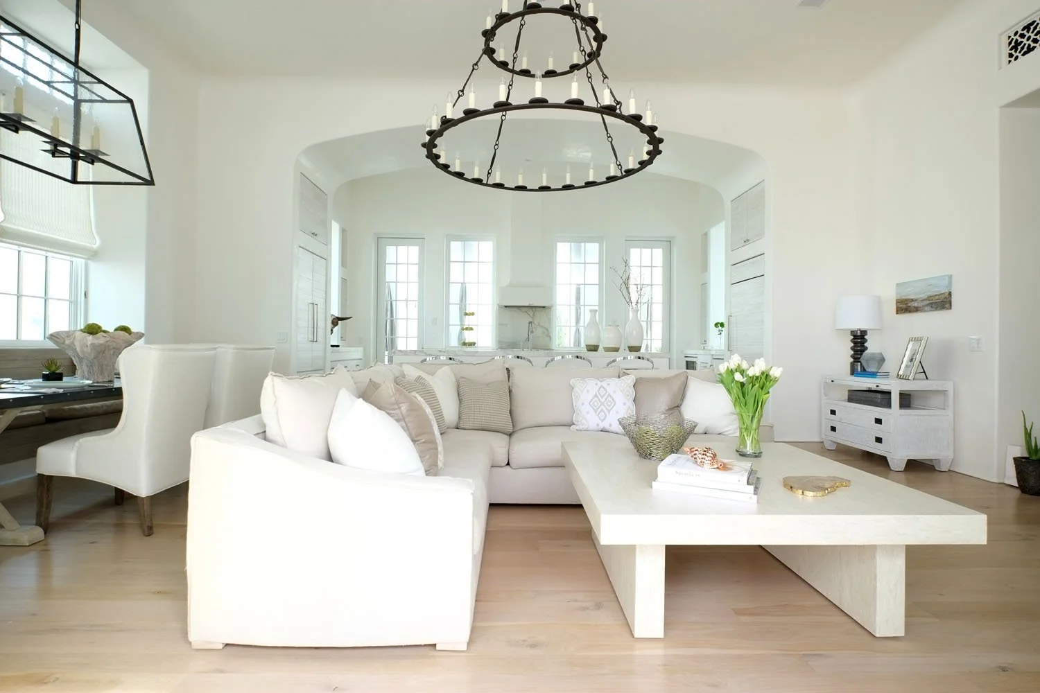 Bright living room with white sofa, matching coffee table with books and flowers, chandelier, and large windows