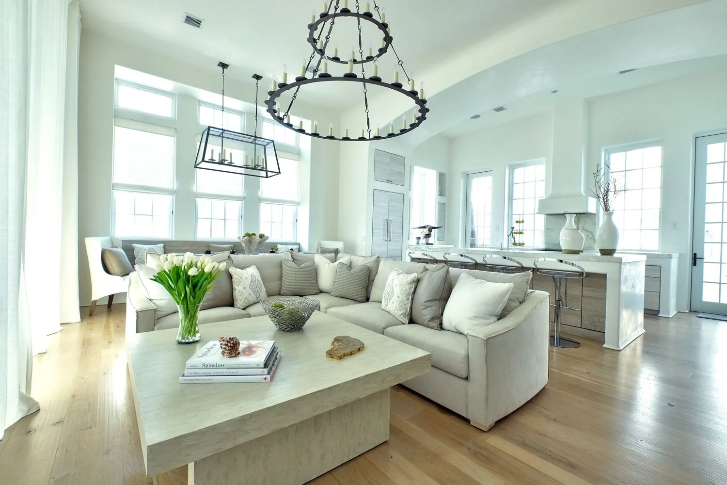 Bright, spacious living room with white walls, large windows, a white sectional sofa with pillows, a wooden coffee table with flowers, books, and decorative objects. There is a large black metal chandelier and a bar area with bar stools, white vases,