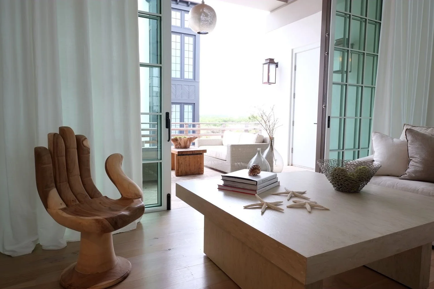 Living room with sliding glass doors opening to an outdoor patio, featuring a wooden armchair, a beige sofa, and a coffee table decorated with starfish, books, and shells, with cushions and vases.
