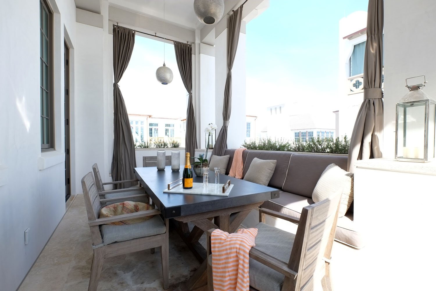 Outdoor patio with a dark rectangular table, six wooden chairs, a bench with cushions, and a tray with champagne and glasses, under large windows with curtains, overlooking buildings and blue sky.