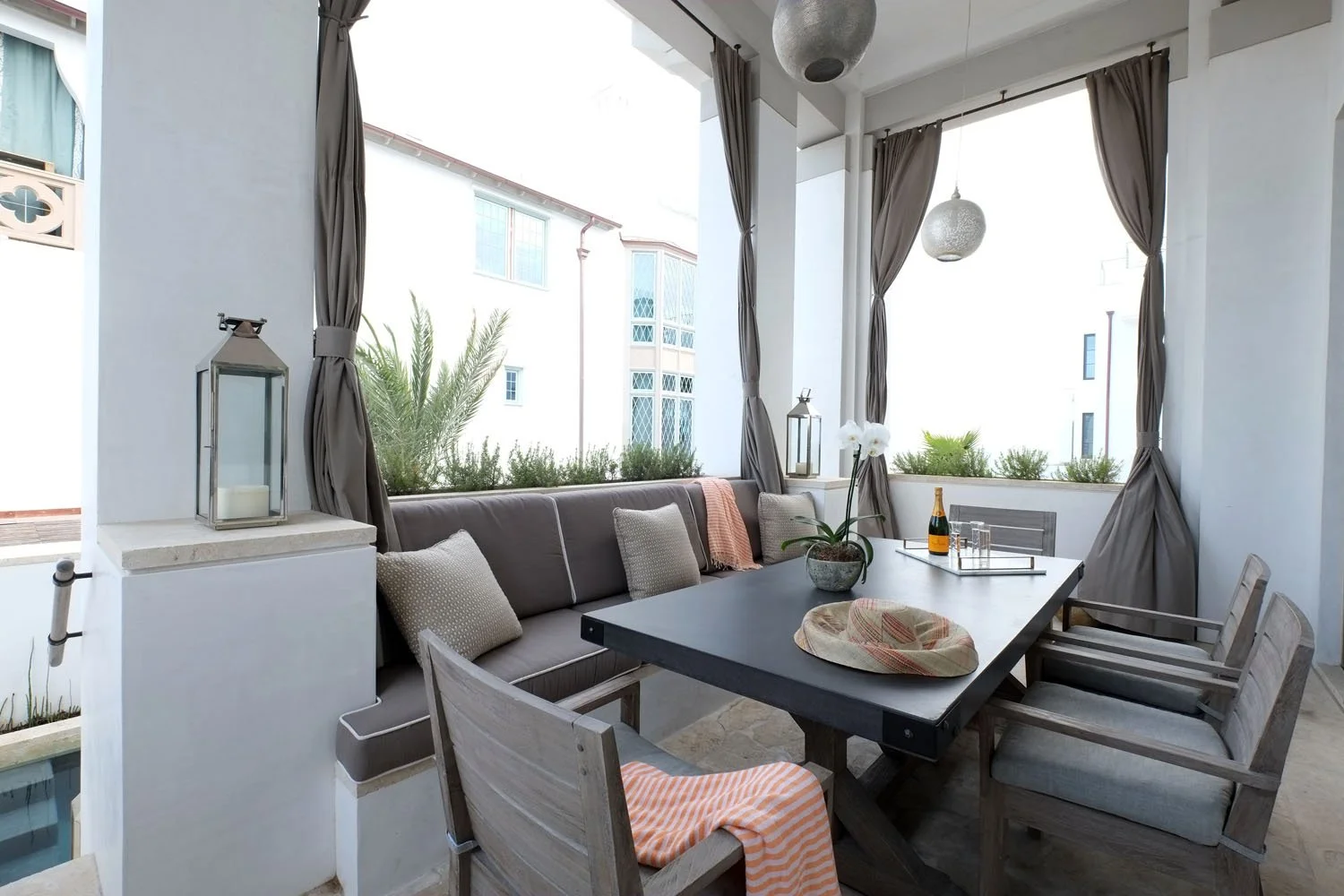 Outdoor patio with dining table, chairs, and decorative lanterns, overlooking a sunny view with modern buildings and greenery.