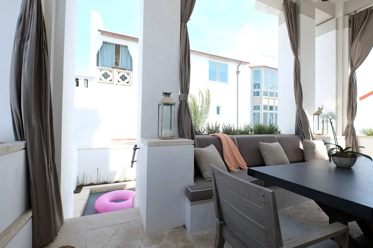 Outdoor covered patio with white walls, grey curtains, a grey outdoor couch with pillows, a black table, a potted orchid, lanterns, and a pink donut float outside on a small pool or hot tub.