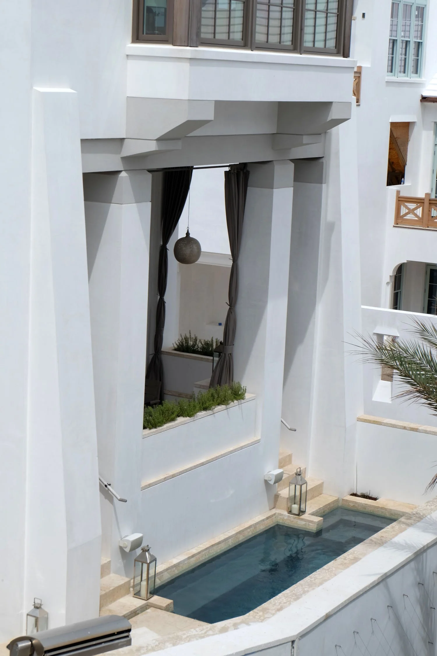 An outdoor courtyard with a rectangular swimming pool, white walls, black curtains, potted plants, lanterns, and a hanging spherical light fixture.