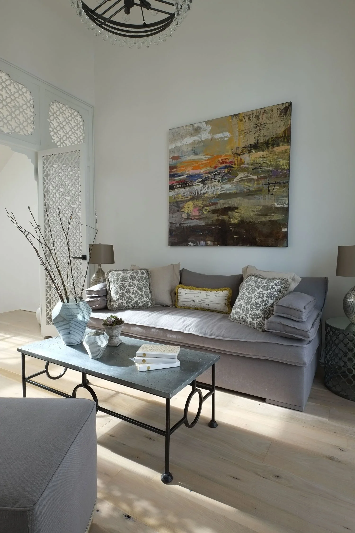 A living room with a gray sofa, decorative pillows, a black metal coffee table with books and vases, a white lattice room divider, table lamps, a large abstract painting, and light wood flooring.