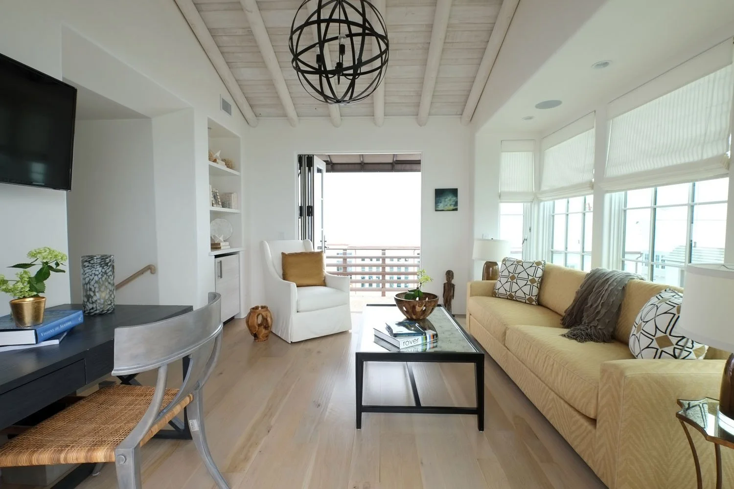 Bright living room with yellow sofa, white armchair with gold pillow, coffee table with plants and books, large windows with blinds, chandelier, and balcony access.