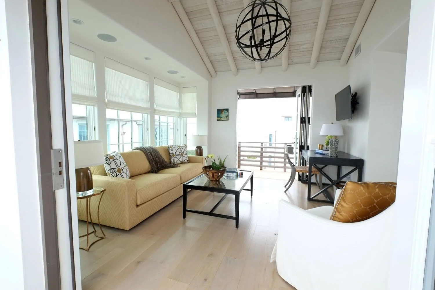 Bright living room with large windows, yellow sofa, black coffee table, white armchair, wall-mounted TV, black console table, and decorative elements in a coastal style.