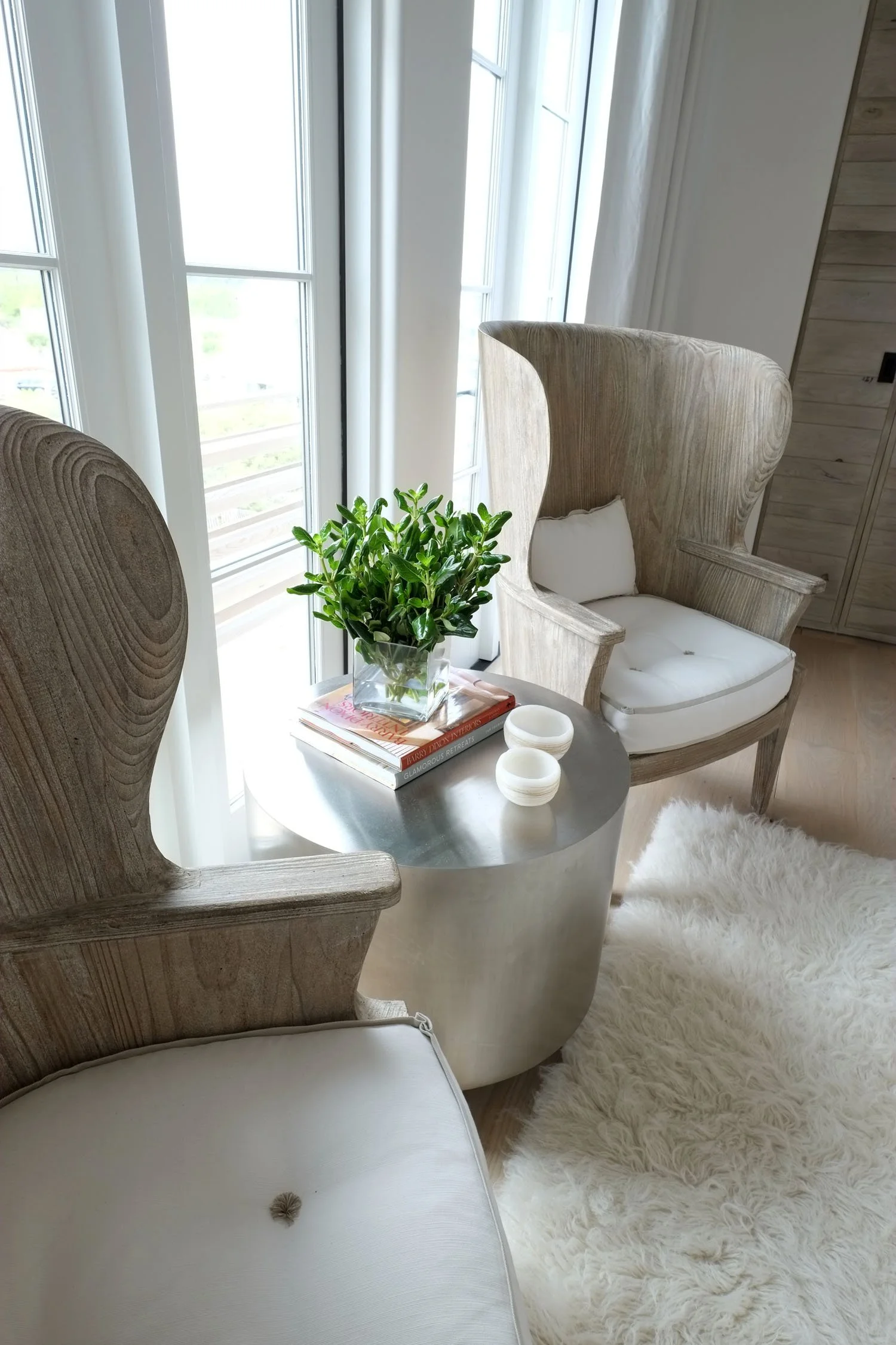 A cozy corner with two wooden armchairs, a small round metallic side table holding a potted plant and books, a fluffy white rug, and a large window with white curtains allowing natural light into the room.