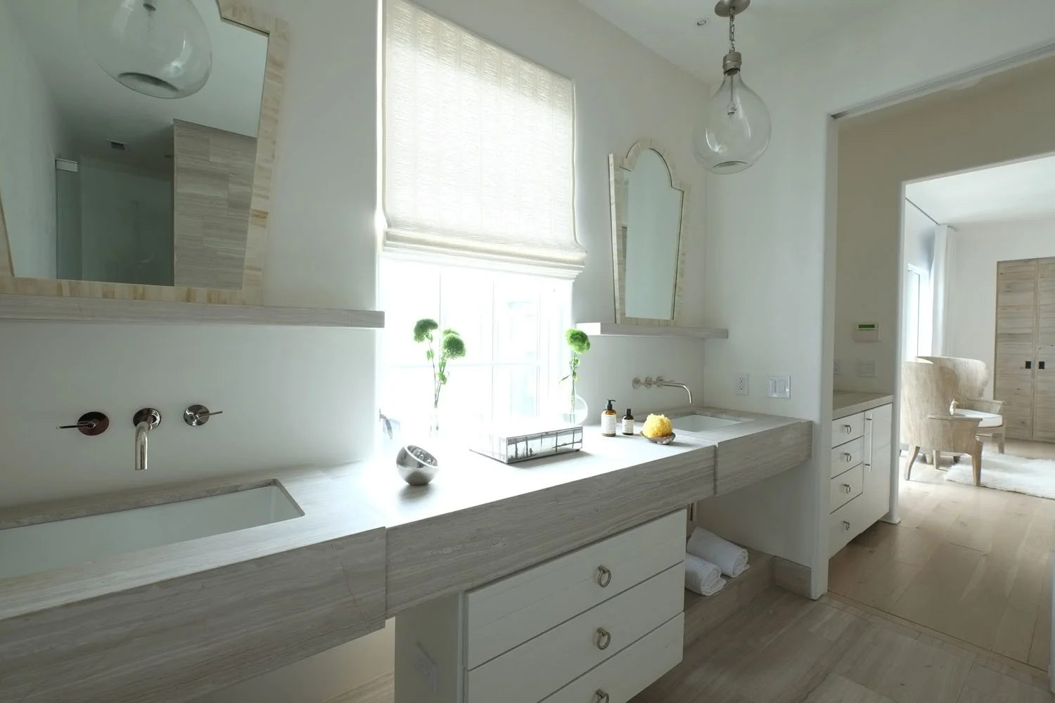 Bright bathroom with white vanity, dual sinks, and a large window with sheer curtains. Decor includes two green plants, a mirror, and minimal accessories. Visible adjoining room with an armchair and wooden wardrobe.