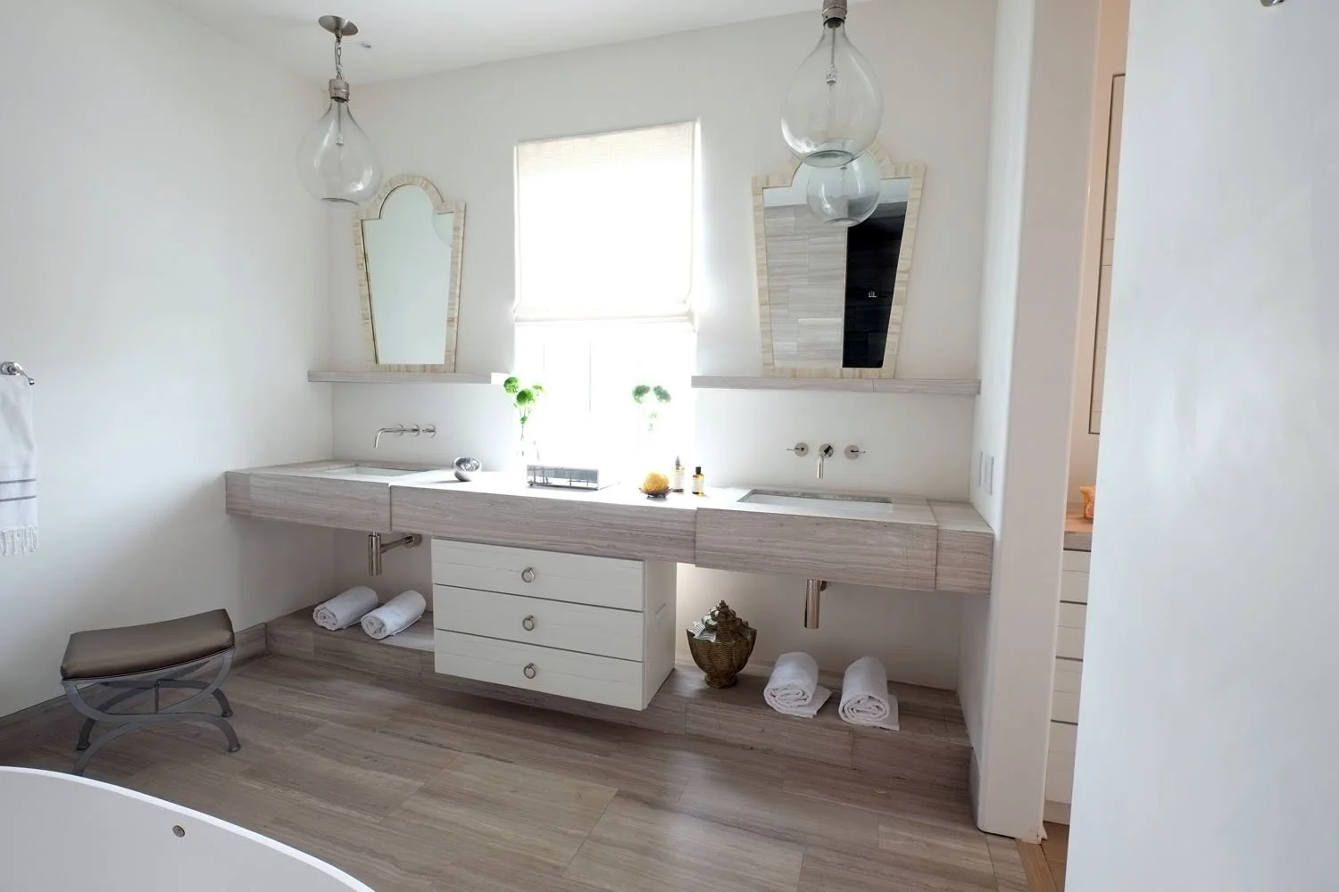 Bright bathroom with double sinks, two mirrors, and a large window in the background. Decor includes white folded towels, green plants, and bottles on the vanity. Light-colored wood flooring and minimalist design.