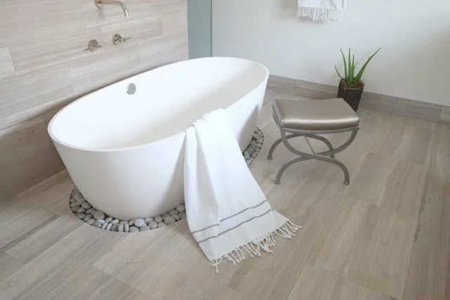 Empty white oval bathtub beside a gray stool with a metal frame, on a light wood floor, with a small potted plant in the corner and a towel hanging on the wall.