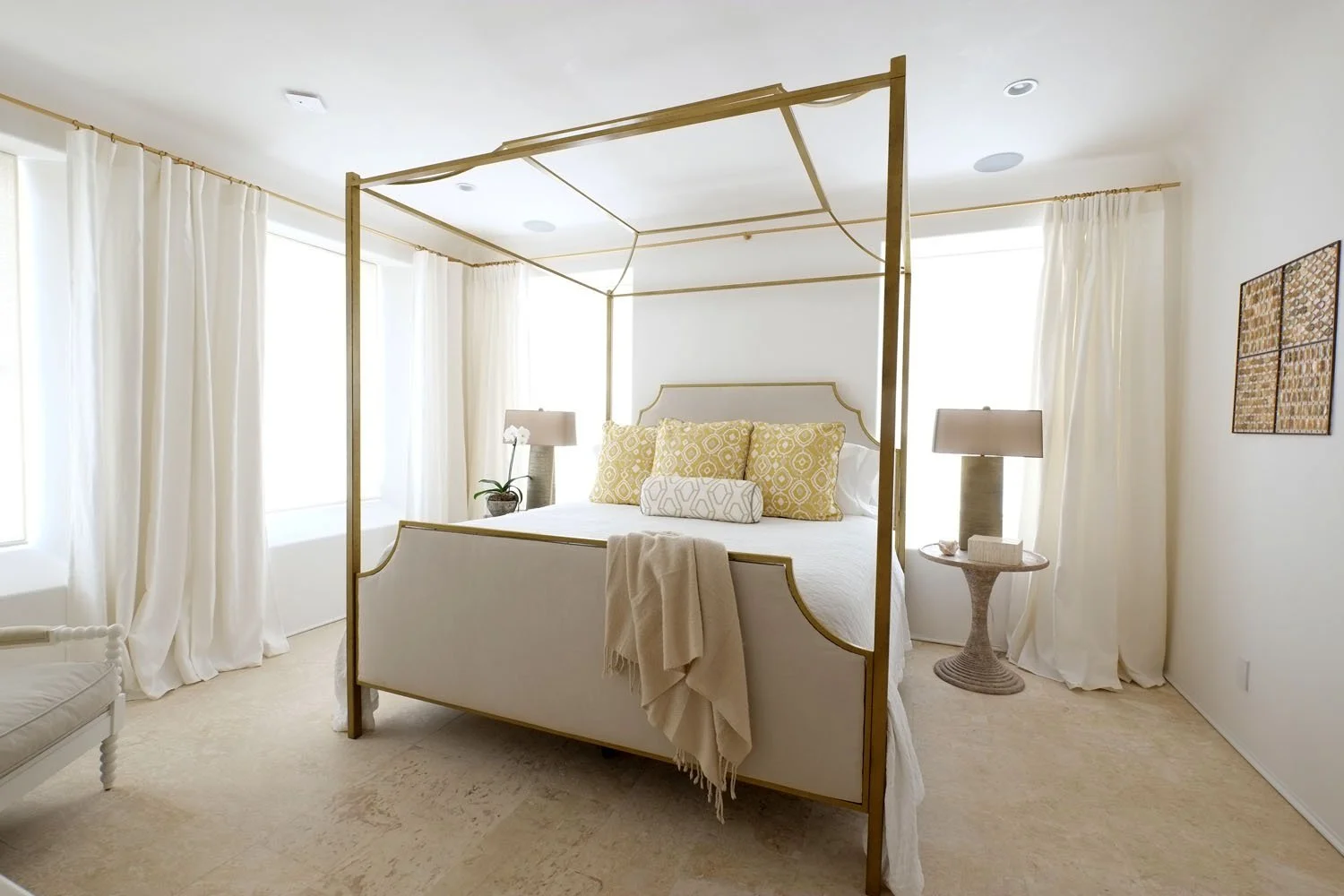 Bright bedroom with white curtains, a four-poster bed with white bedding and yellow pillows, two bedside tables with lamps, and artwork on the wall.