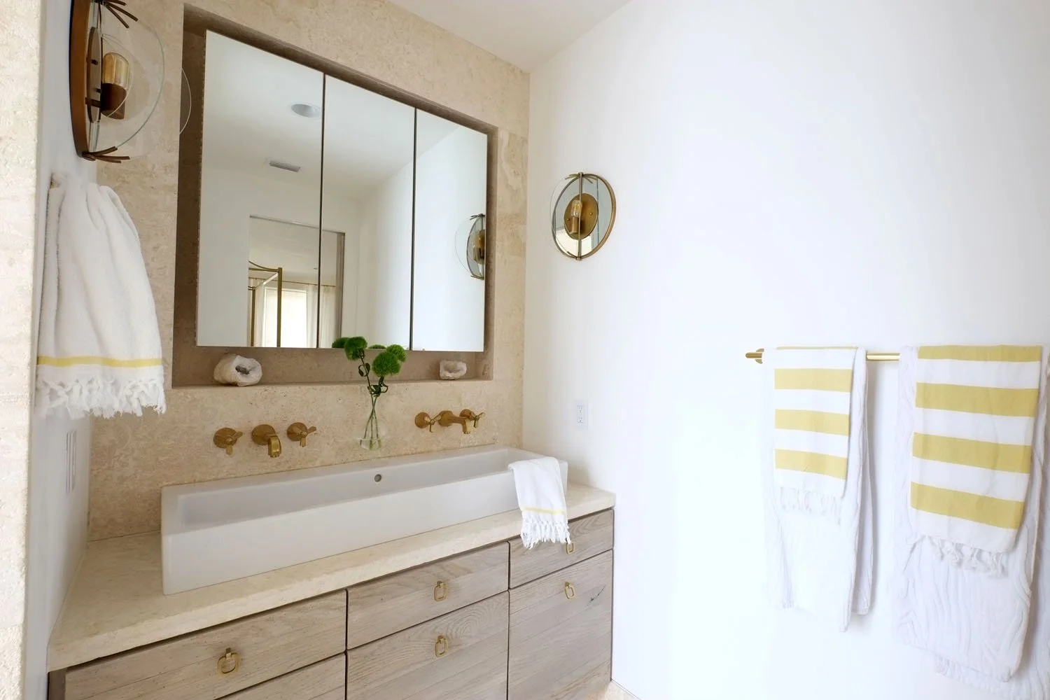 A modern bathroom featuring a large mirror, a beige marble countertop with a rectangular white sink, gold fixtures, and yellow striped towels hanging on a wall hook.