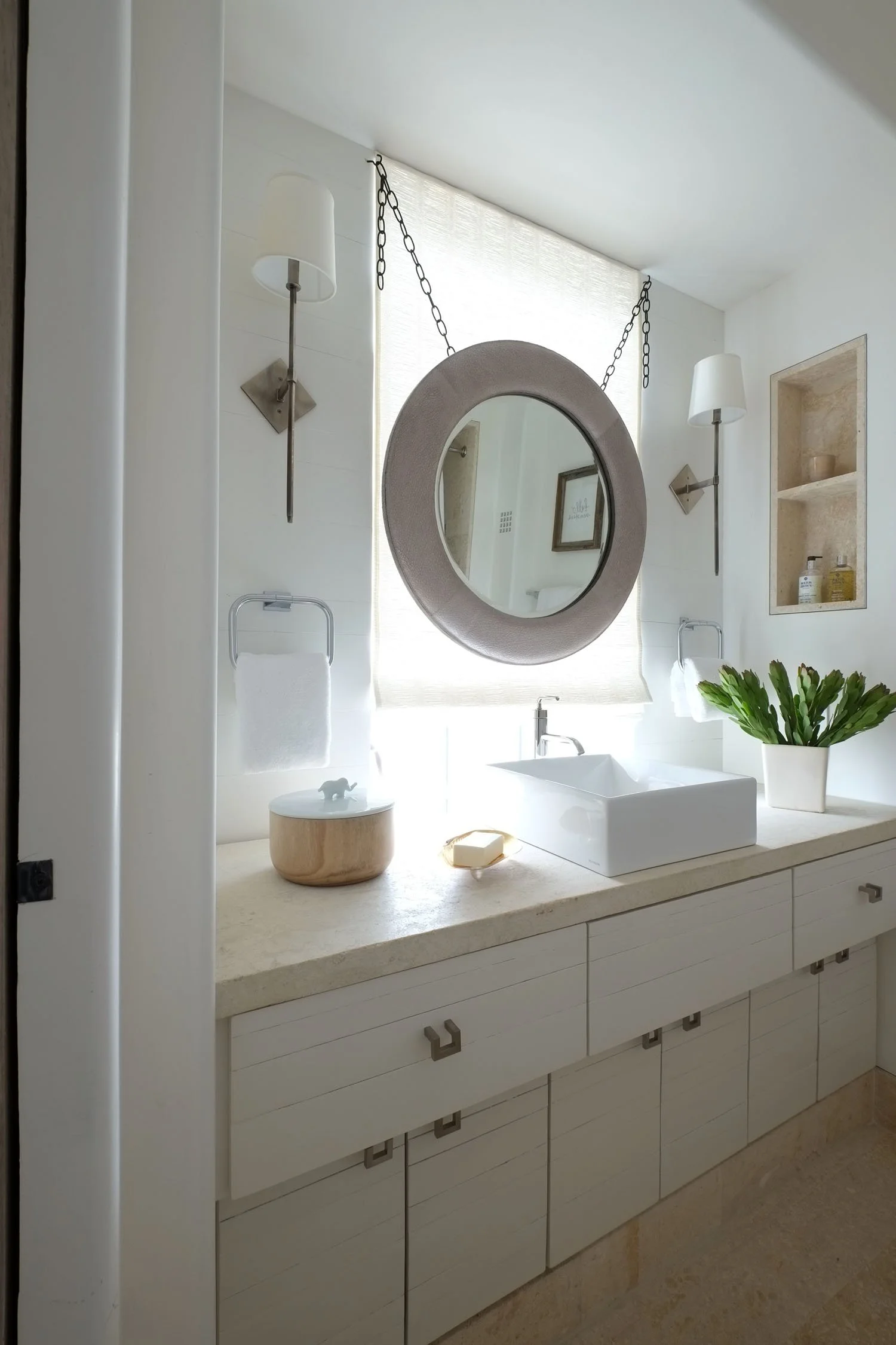 Modern bathroom with a beige countertop, white sink, two wall-mounted lights, a round mirror hanging from chains, a potted plant, and built-in shelves with toiletries.
