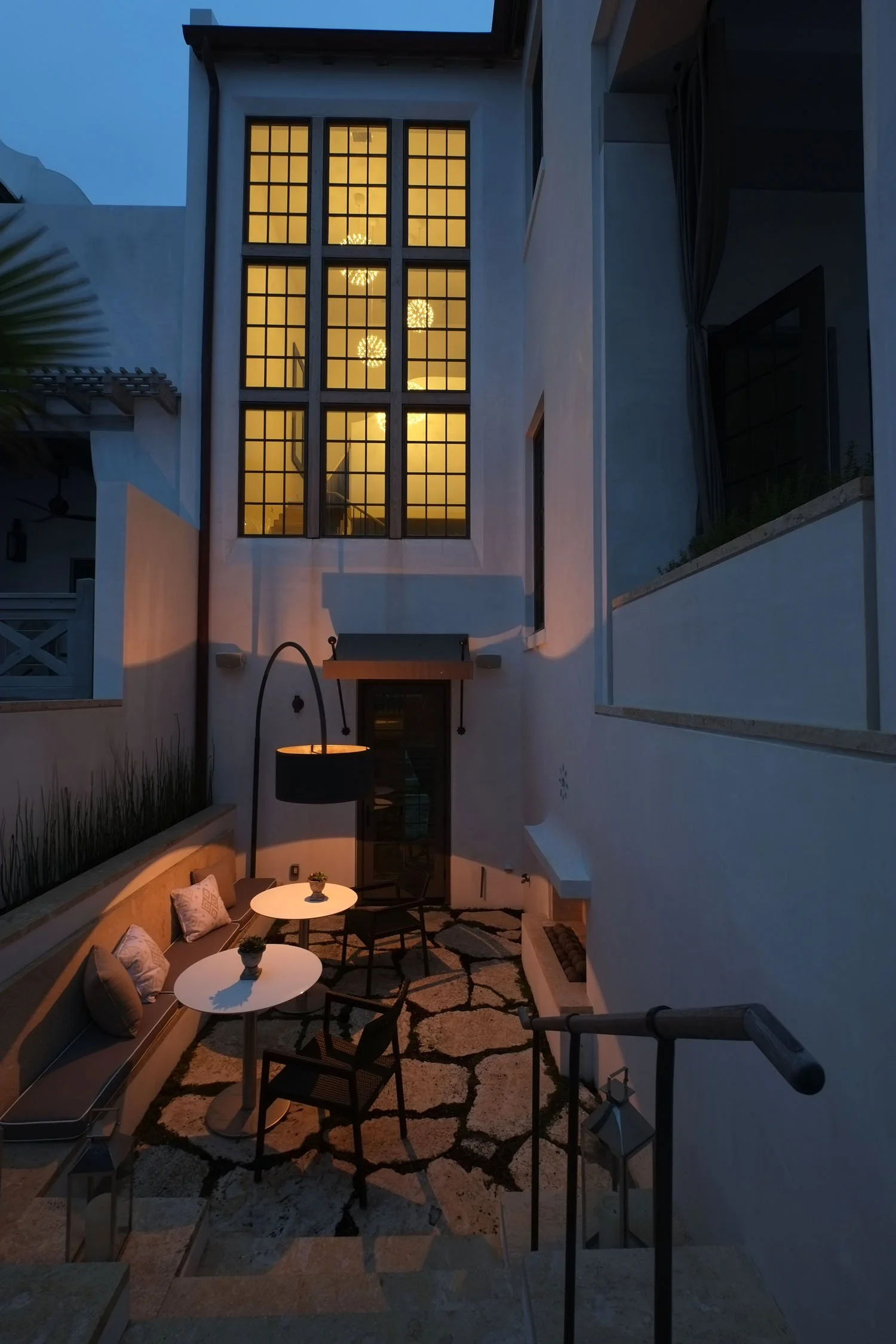 A cozy outdoor patio enclosed by white walls, with a built-in bench, two white tables, and chairs, illuminated by a large lamp. Large windows above reveal a chandelier inside the house, with lighting creating a warm ambiance at dusk.