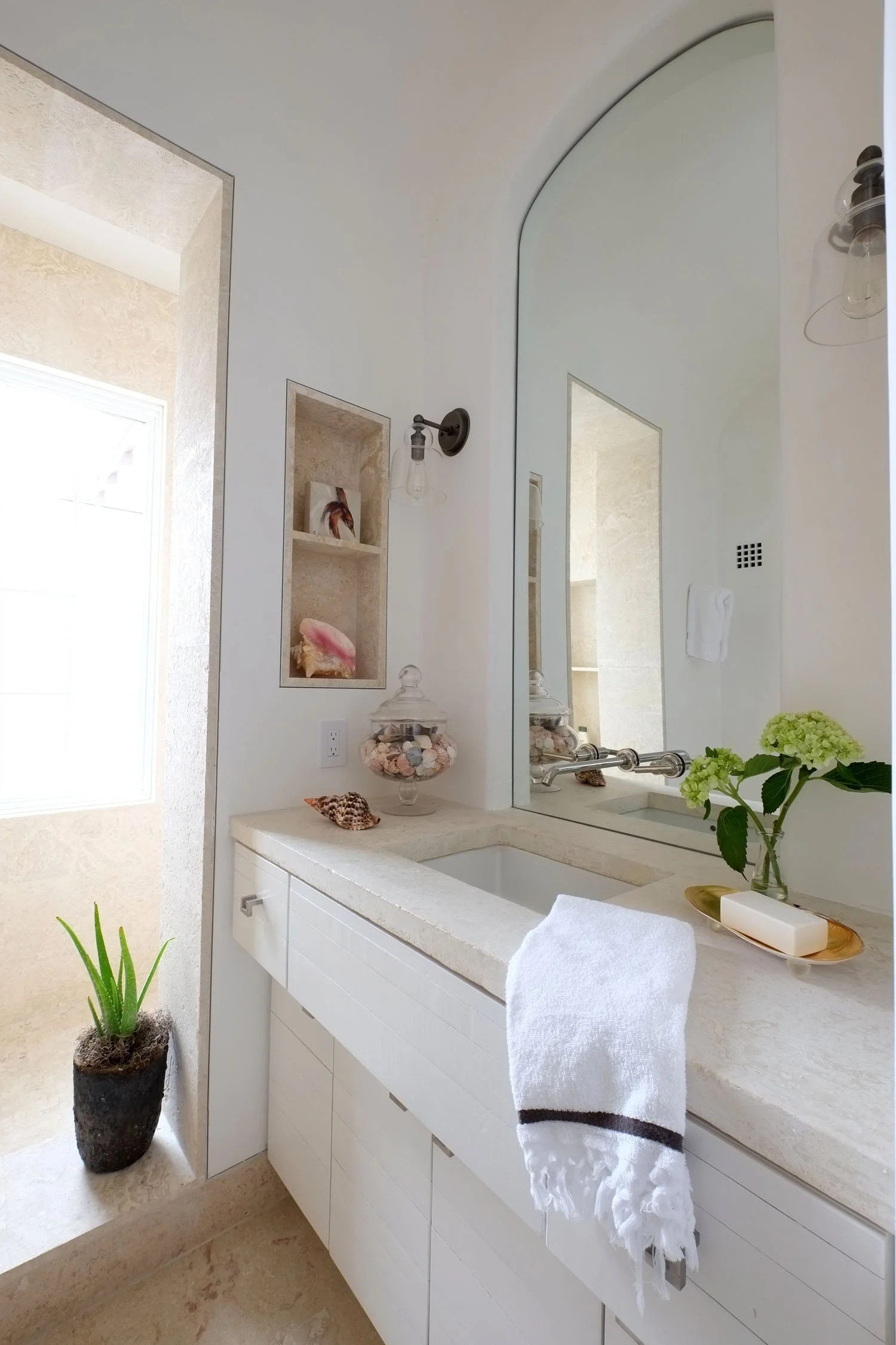 A bathroom with a white vanity, a large mirror, a small sink, decorative shells, flowers, and natural light from a window.