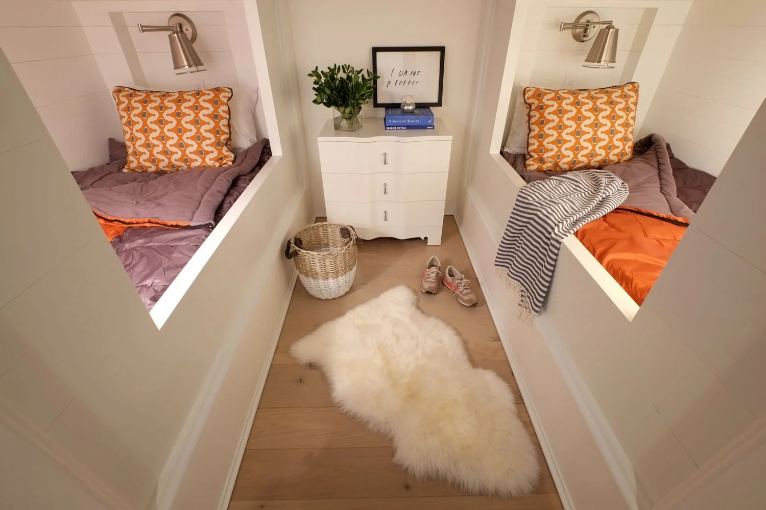 Two enclosed beds with orange and gray bedding and geometric pillows, a small white dresser with a plant, framed art, and books in between, a fluffy white rug, and sneakers on a wood floor.