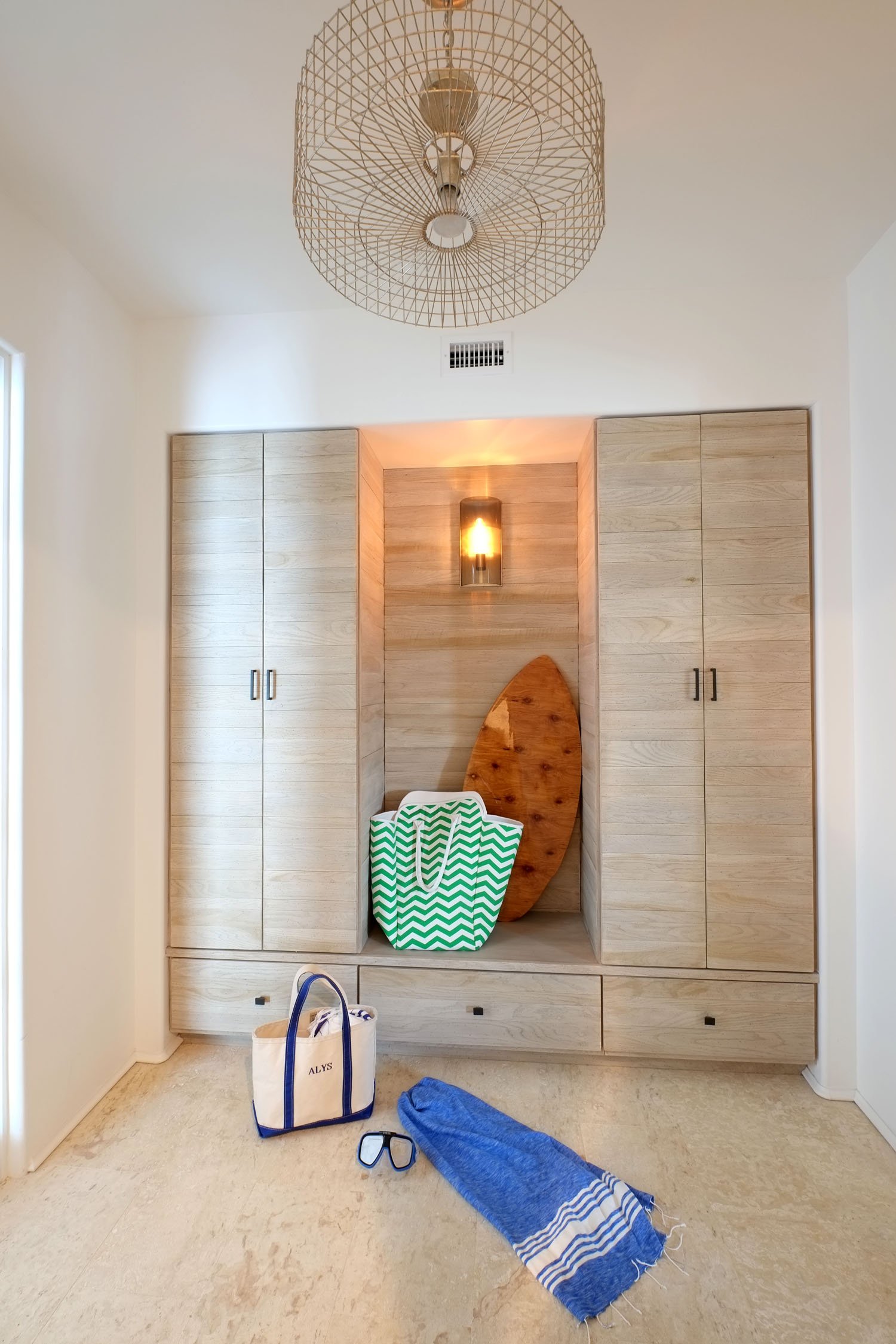 Entryway with built-in wooden storage cabinet, a wooden surfboard, a green and white striped bag, a blue towel, a tote bag, and sunglasses on a beige floor. Overhead lighting and wall-mounted light fixture.