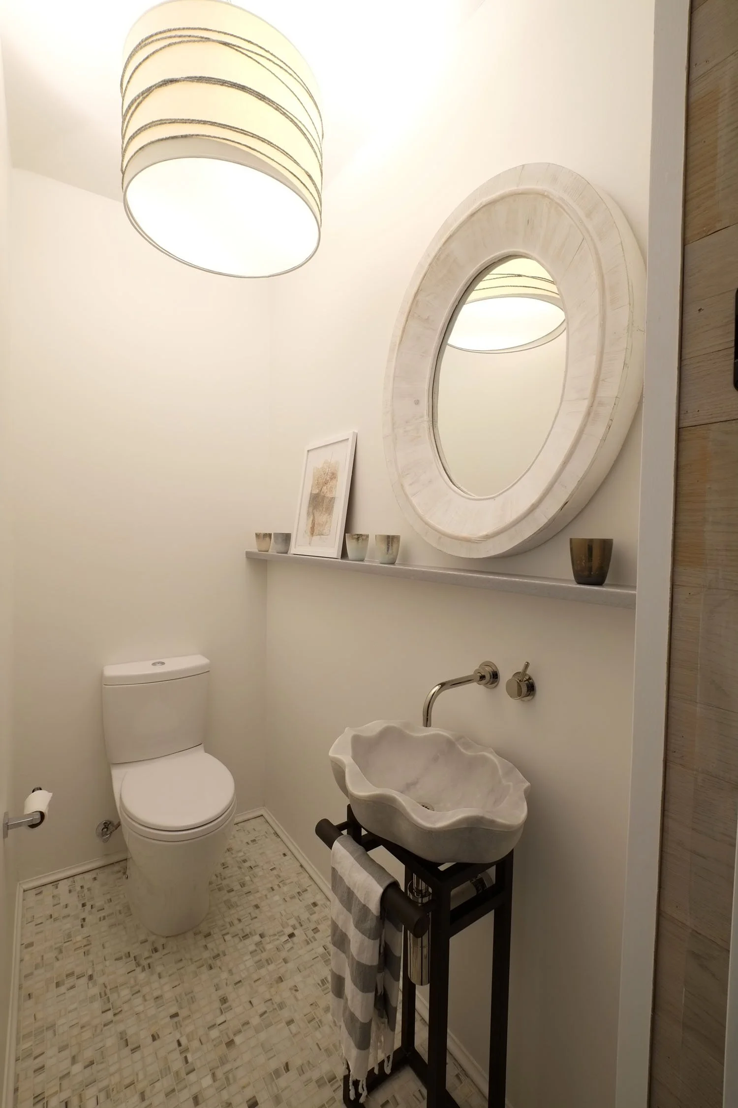 A small bathroom with a white toilet, a stone sink on a black stand with a towel hanging, a circular mirror, a shelf with framed picture and small cups, modern ceiling light, and wood-paneled wall on the right.