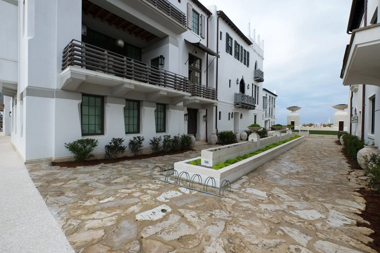 View of a narrow cobblestone pathway between white Mediterranean-style buildings with balconies, potted plants, and landscaped garden beds. In the distance, there are two decorative statues with large bowls on top.