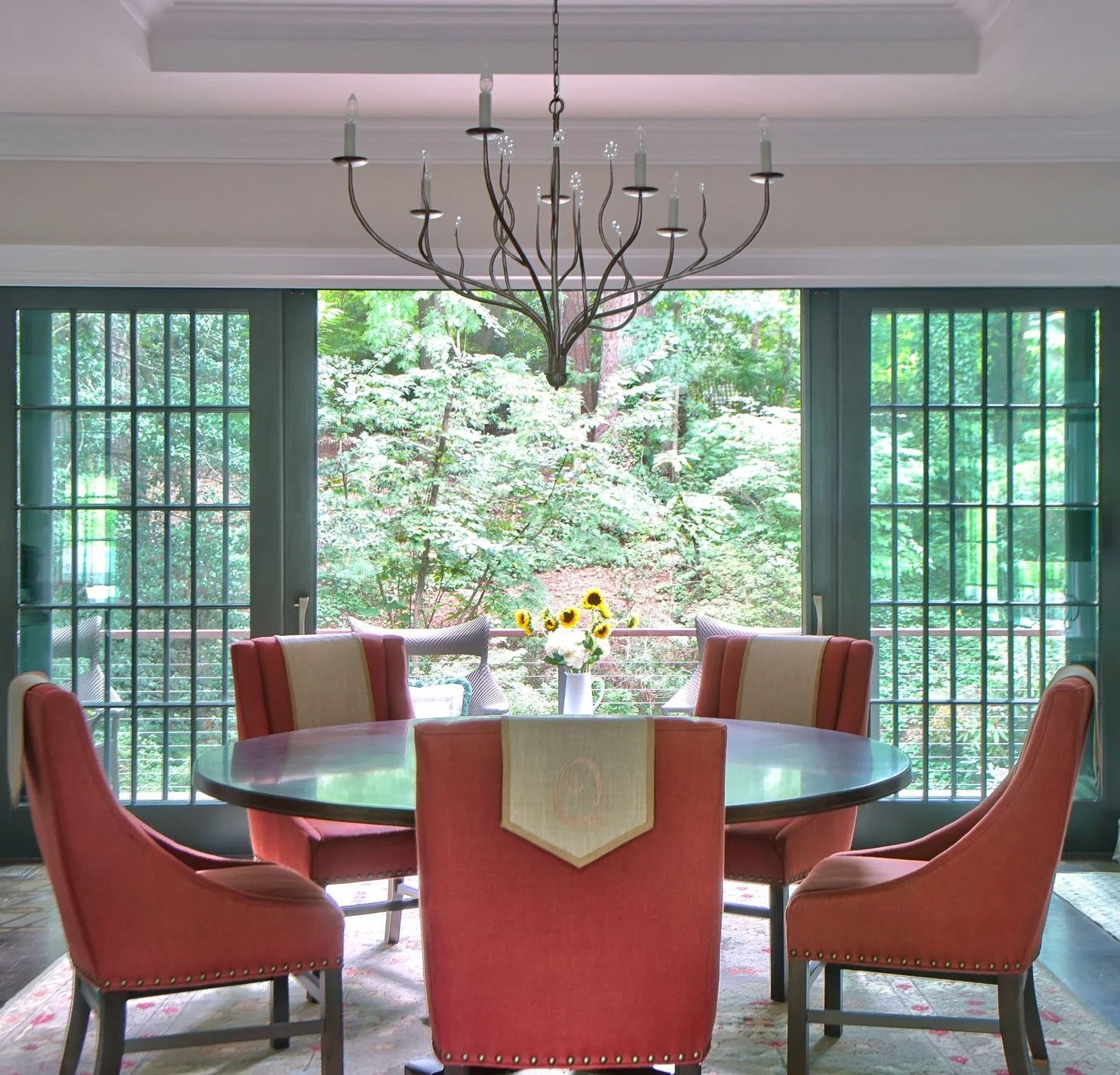 A round dining table with six pink upholstered chairs is situated in front of large sliding glass doors that open to a lush green forest. A white vase with sunflowers is on the table, and a black wrought iron chandelier hangs from the ceiling.